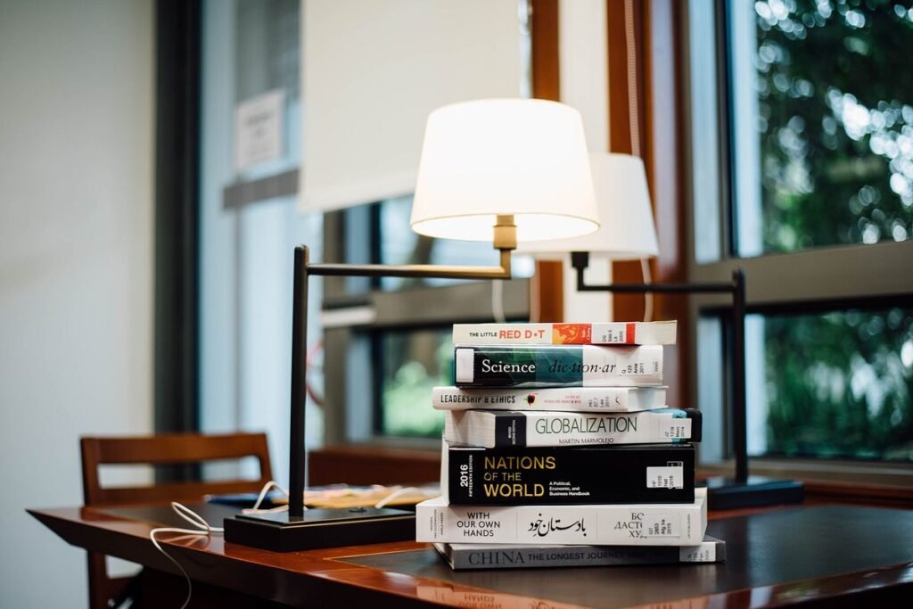 Pile de livres posée sur un bureau en bois près d'une lampe, devant une fenêtre